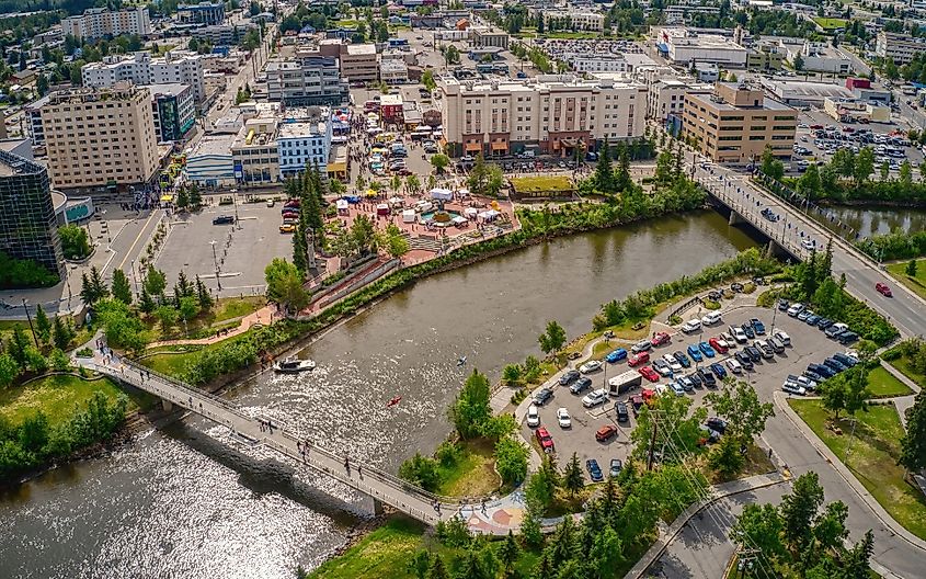 Aerial view of a lively cityscape featuring a river with two pedestrian bridges, leafy parks, parked cars, and surrounding urban buildings under a bright sky.