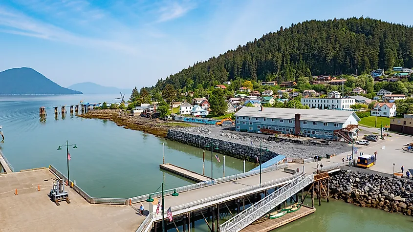 A scenic coastal town with a small harbor, surrounded by lush green hills and colorful houses. A clear blue sky and calm water create a peaceful atmosphere.